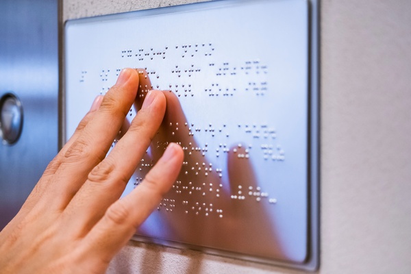An individual using their fingers to read the tactile dots of a silver braille sign mounted on a wall.