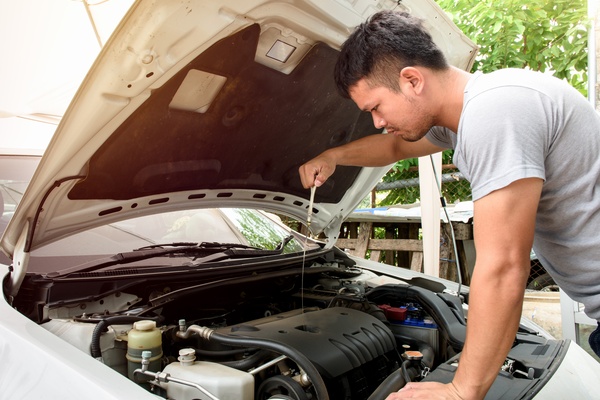 A man wearing a gray t-shirt standing in front of his car with the hood open while checking the oil level with the dipstick.