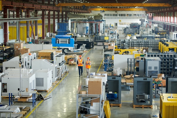 A large production warehouse filled with machinery. Two workers are walking between the equipment in safety gear.
