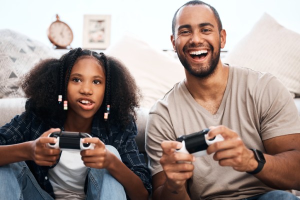 A smiling teen girl and her father sit on the floor in front of a sofa. They hold controllers while playing a video game.