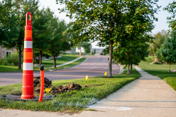 An orange traffic cone sits next to a pile of dirt over a hole in the ground between a residential street and sidewalk.