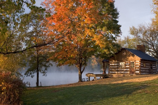 A wood cabin sits next to a lake. It's fall, and leaves on the trees have changed color. There's fog coming off the lake.