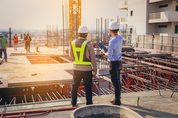 Two men wearing white hard hats are standing, looking at a construction site. One man is in business casual clothing.