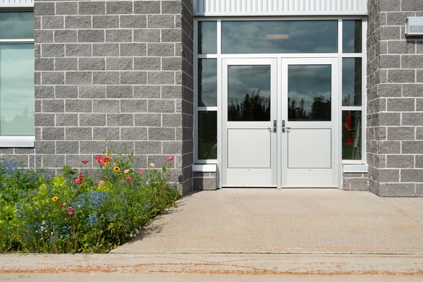 Outside of a brick building with two steel doors. Along the path to the door, flowers of different colors are growing.