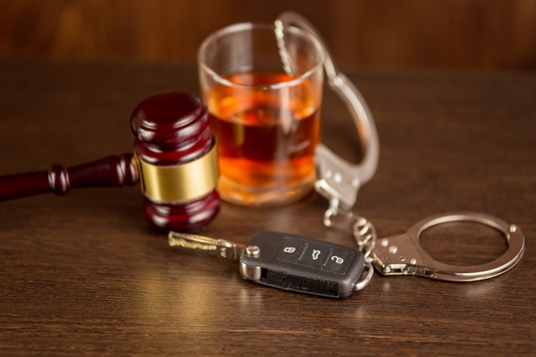 A set of car keys rests on a brown wooden table next to a glass, a pair of handcuffs, and a wooden gavel.
