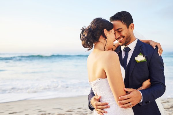 A young couple wearing a wedding suit and dress are embracing each other on a sandy beach. The ocean waves behind them.
