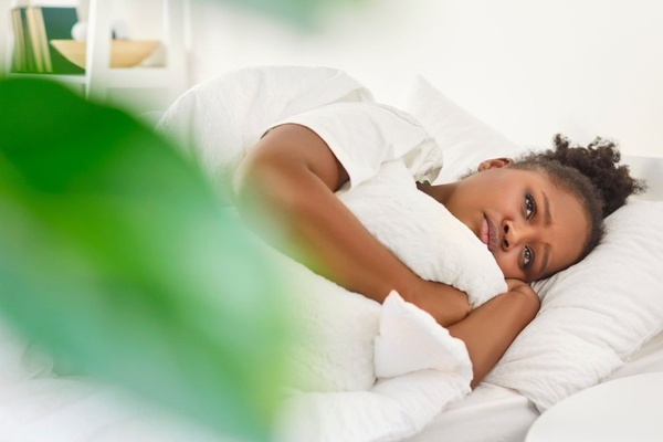 A Black woman laying awake in her bed in the morning. Her eyes are heavy and restless, and her linens are white.