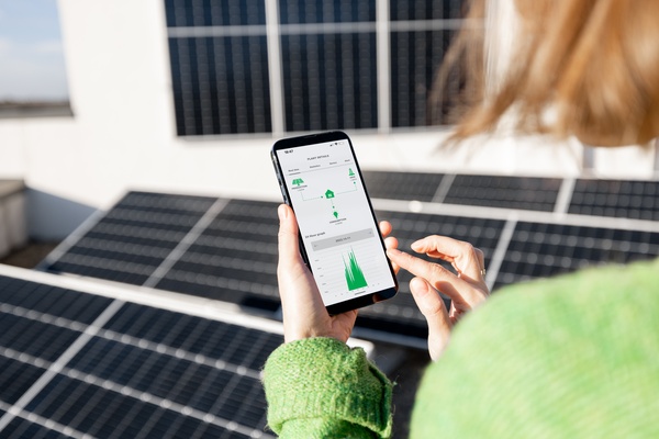 A person in a green sweater looks at a green graph on her smartphone. She stands on a roof fitted with solar panels.