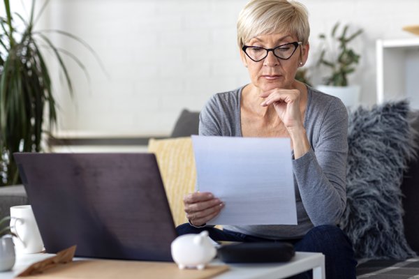 An older woman with short blonde hair and glasses sits indoors, reading a sheet of paper with a laptop in front of her.