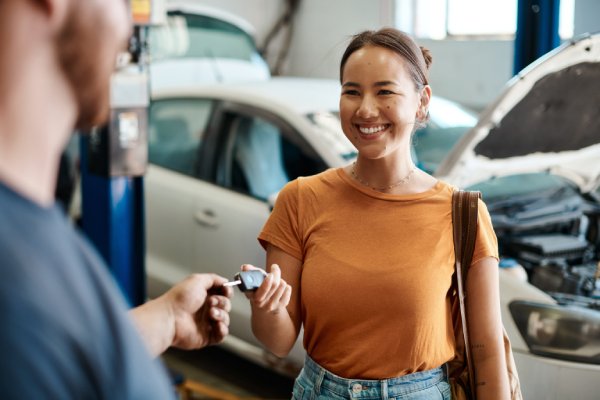 A woman handing her key over to a mechanic in an auto shop. Behind her is a car with the hood lifted.