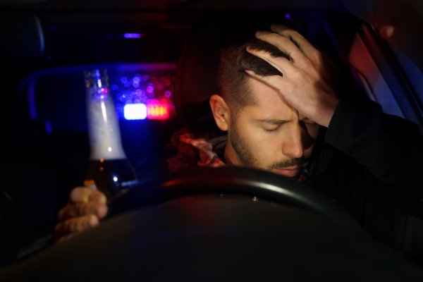 A man sits in the driver's seat of a car while holding a glass bottle. Police lights flash in the background.