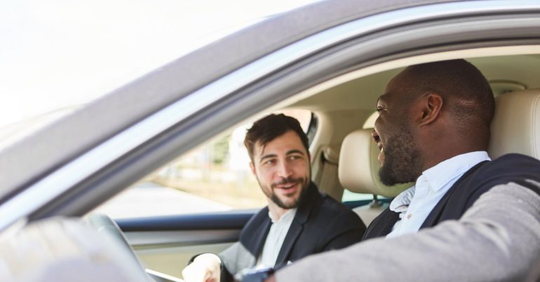 Two coworkers sit in a car, talking and laughing. They carpool as the commute to work to reduce their carbon footprint.