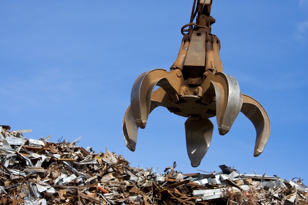 A rusted crane claw dropping down over the top of a pile of rubbish. The pile contains rusty scraps and cut up metal.