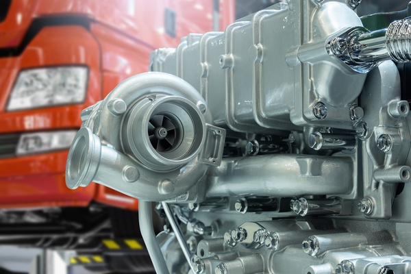 A close-up view shows a silver metal turbo attached to an engine. A red truck appears blurry behind the engine.