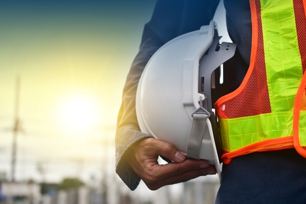 A technician holding a white hard hat with the sunlight in the background. They also have a construction vest on.