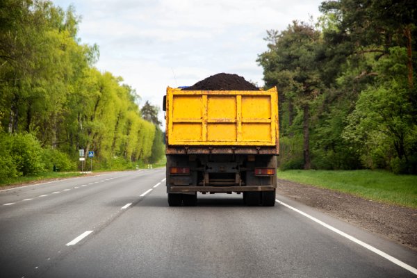 The back view of a yellow dump truck transporting black soil along an asphalt road in a wooded area of town.