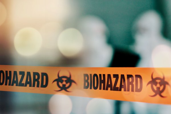 An orange piece of tape sectioning off an area. The tape says “Biohazard” and two people wearing full PPE are nearby.