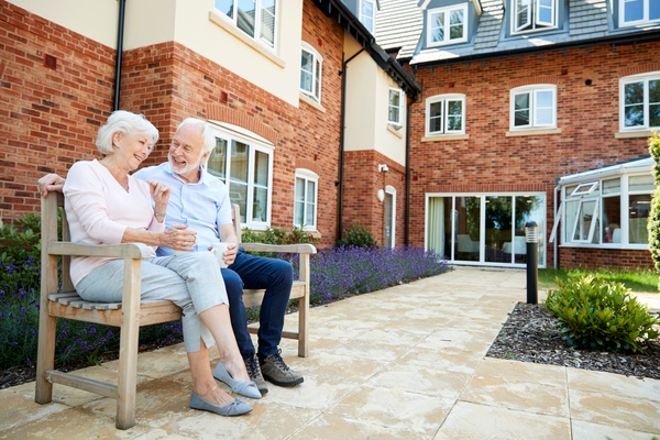 Two older people are sitting next to each other on a wooden bench. They are both smiling and the bench is outside.