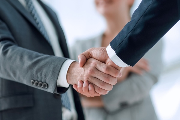 Two professionally dressed people shake hands. Another person in a business suit stands nearby and smiles.