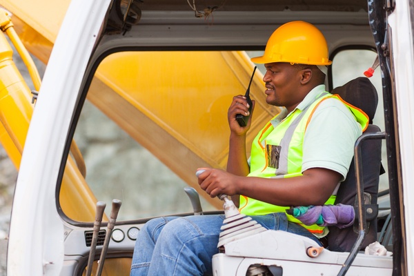 A construction worker sits in the cab of a heavy-duty machine. He is wearing high-vis gear and talking into a handheld radio.