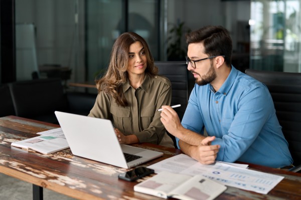 Two business professionals sit at a long wooden desk. A laptop and various papers are on the surface.