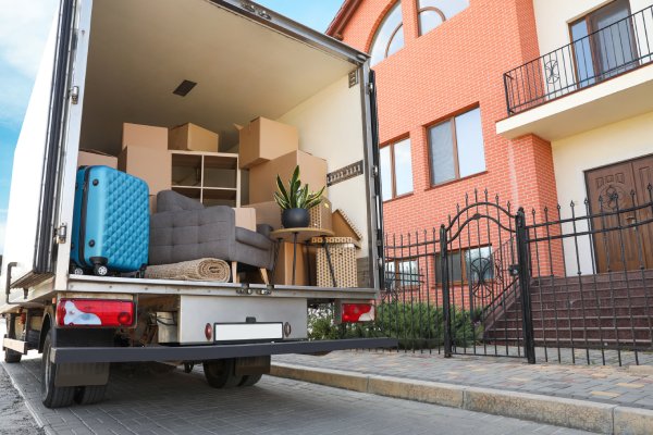 A moving truck sits on a street in front of an apartment building. The truck is full of boxes, luggage, and furniture.