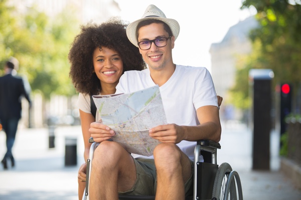 A happy couple posing together while outside on a vacation. The man is in a wheelchair, holding a map.