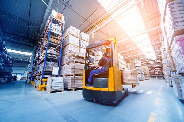 A forklift loading packages from storage shelves onto the prongs. The storage is in a large, clean warehouse.