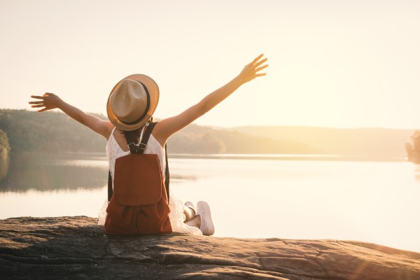 A woman sits on a rock that overlooks a lake at sunset. She wears a backpack and stretches out her arms to embrace the scenery.