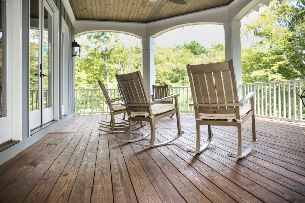 Four cream-colored rocking chairs sit on a large front porch with a white railing. The porch has a wooden floor.