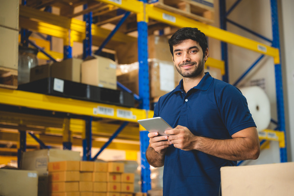 A man standing in a warehouse with large storage units behind him. He has a small tablet in his hands.