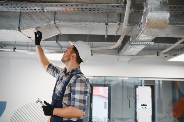 A person wearing a white hard hat is holding a tool and reaching up to work on the ductwork of an HVAC system.