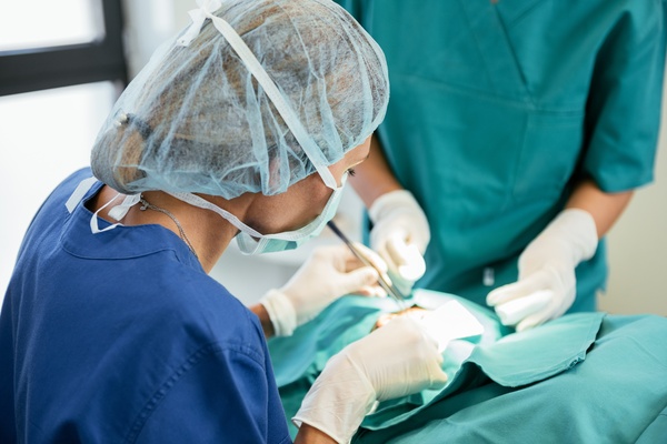 A female medical professional using a surgical tool on a patient. She's wearing a mask, hair net, and gloves.