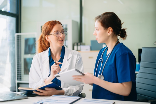 A doctor talking to a nurse inside an office. The doctor is holding a notebook and the nurse has a tablet.