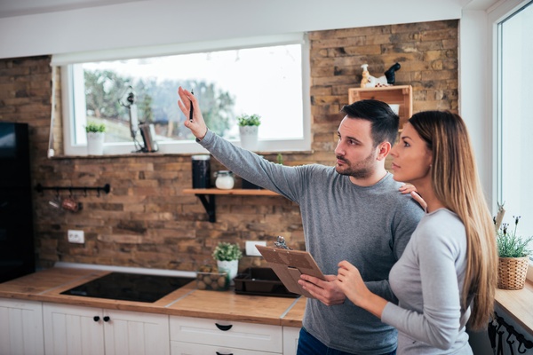 A man and a woman are standing in a kitchen. The man is holding a clipboard and gesturing to something out of frame.