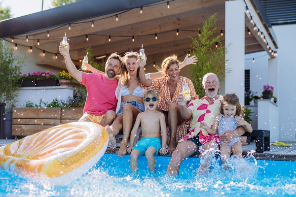 A family of six, including two toddlers, sitting by the edge of a pool, dipping their feet, cheering, and holding drinks up.