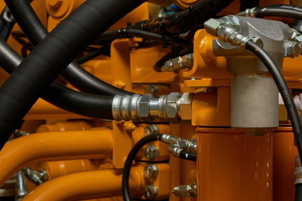 A close-up of a section of industrial hydraulic machinery. The metal pipes are orange, the tubing black, and the fittings silver.