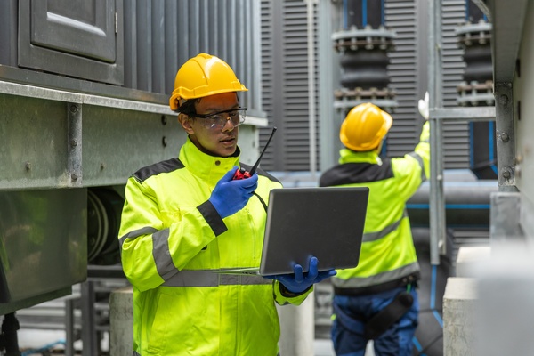 Two workers in safety gear: one uses a laptop and a walkie-talkie, the other inspects equipment in an industrial setting.