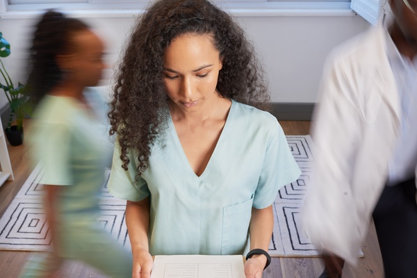 A woman wearing scrubs is looking down at a clipboard she is holding as other healthcare professionals rush behind her.
