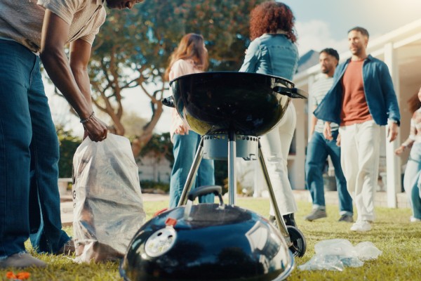 A group of people are in a yard. One person is opening a bag next to a grill. There are trees in the background.
