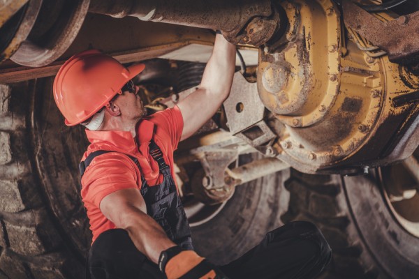 A man wearing a hard hat is crouched beneath heavy mining equipment. The equipment has dirt on the tires and metal.