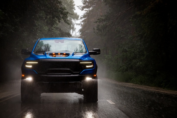 A blue pick-up truck with its headlights on, driving on a highway between rows of tall trees on a rainy day.