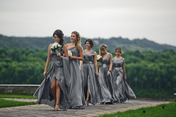 Five bridesmaids in grey dresses walk outdoors with white bouquets, set against a green landscape and cloudy sky.