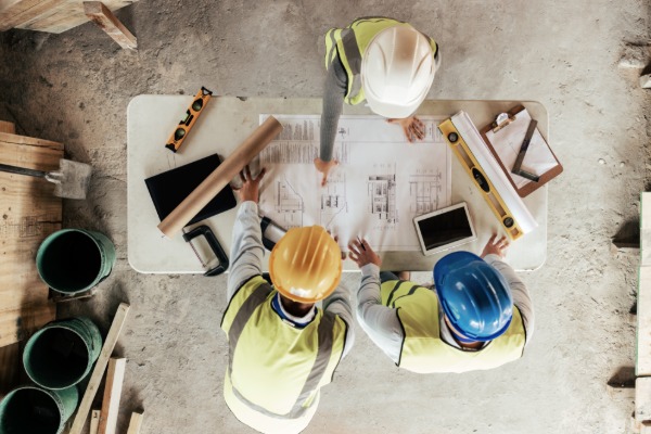 Three construction workers reviewing the schematics for a project. All of them are wearing different colored hard hats.