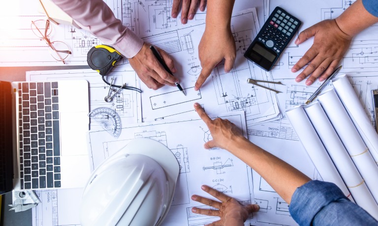 A bird's eye view of people pointing at blueprints on a table scattered with design instruments and tools.