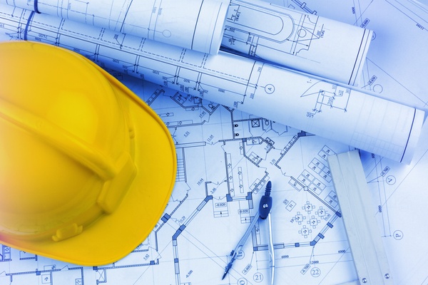 A yellow, hard hat sits on top of rolled up architectural blueprints, next to a drawing compass and a gray level.