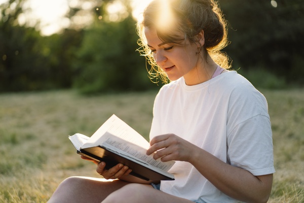 A young woman wearing a white T-shirt and shorts sits outside in a grassy area reading a book. She holds a page in one hand.