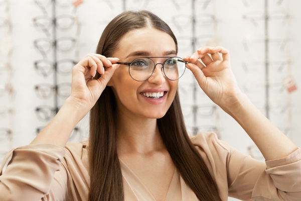 A woman trying on a pair of glasses while in a glasses store. There are multiple rows of glasses on stands behind her.