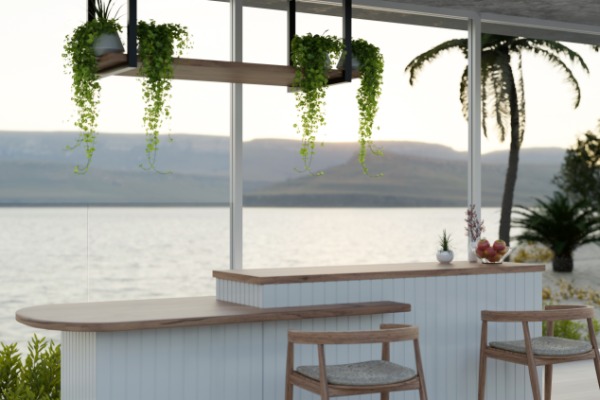 An indoor bar with wooden countertops looks out windows to a large body of water. Indoor plants hang over the bar.