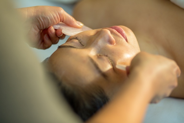 A person laying on their back with their eyes closed as a beauty technician smooths down their cheeks with two gua sha tools.